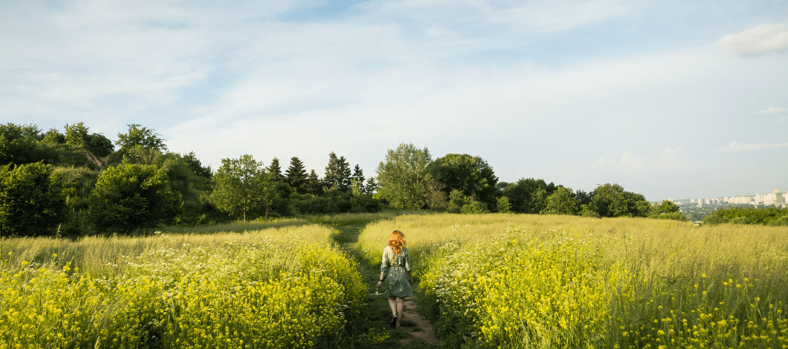 mulher a passear por um caminho que atravessa um campo agrícola florido, com árvores ao fundo