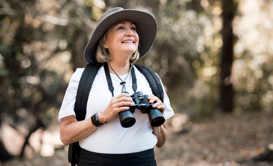 mulher de meia-idade sorridente a segurar nos binóculos que tem ao pescoço. O fundo desfocado parece um bosque no verão